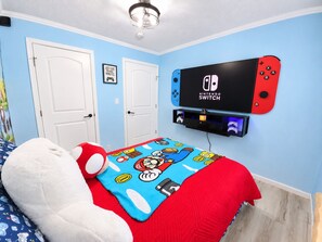 A bedroom with a Nintendo Switch mounted on the wall, a bed with a Mario-themed blanket, and a plush toy.