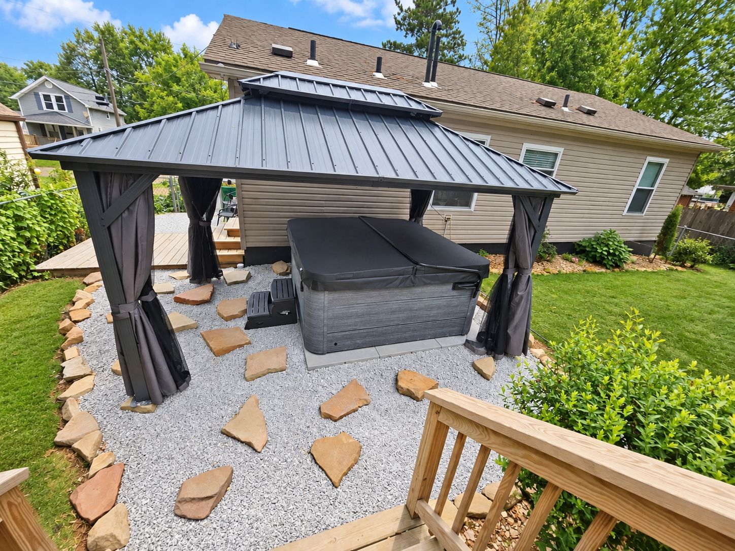 A covered outdoor seating area with a bench and a table, surrounded by a gravel path and a wooden deck.