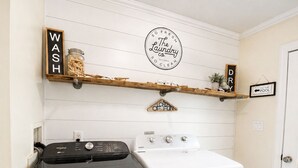 Un'area lavanderia con una lavatrice Whirlpool e un'asciugatrice Maytag, un cartello che recita 'The Laundry Co. Self-Serve So Clean' ed scaffali di legno con articoli per la lavanderia.