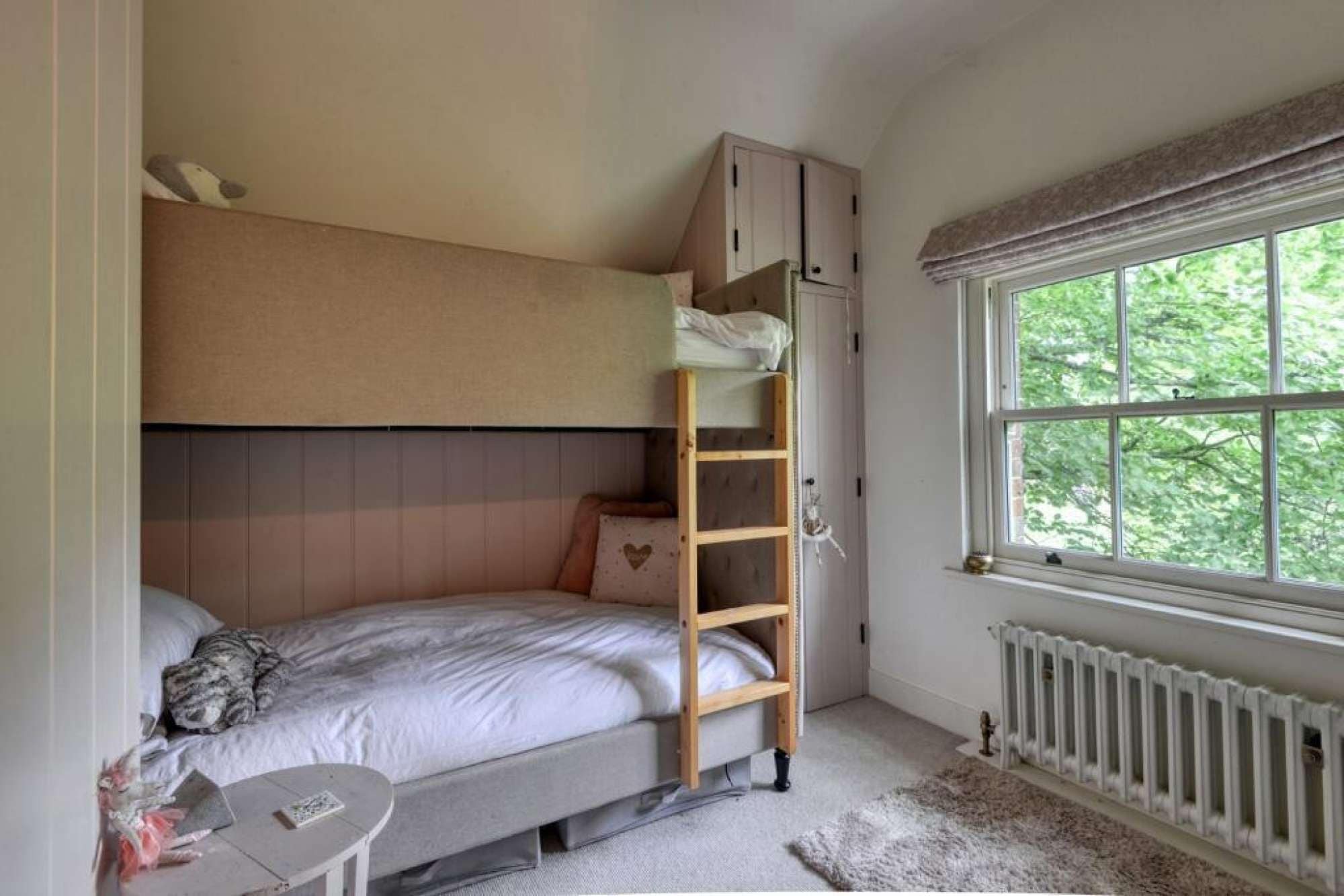 A bedroom with a bunk bed, a ladder, a window with a view of greenery, and a radiator.