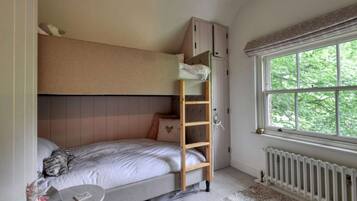 A bedroom with a bunk bed, a ladder, a window with a view of greenery, and a radiator.