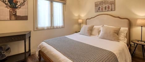 A bedroom with a bed, a bedside table, a lamp, a window with curtains, and artwork on the walls.