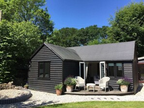 A black wooden house with a porch, white chairs, and potted plants. - The Cabin – A Boutique Retreat in the New Forest (Brockenhurst)