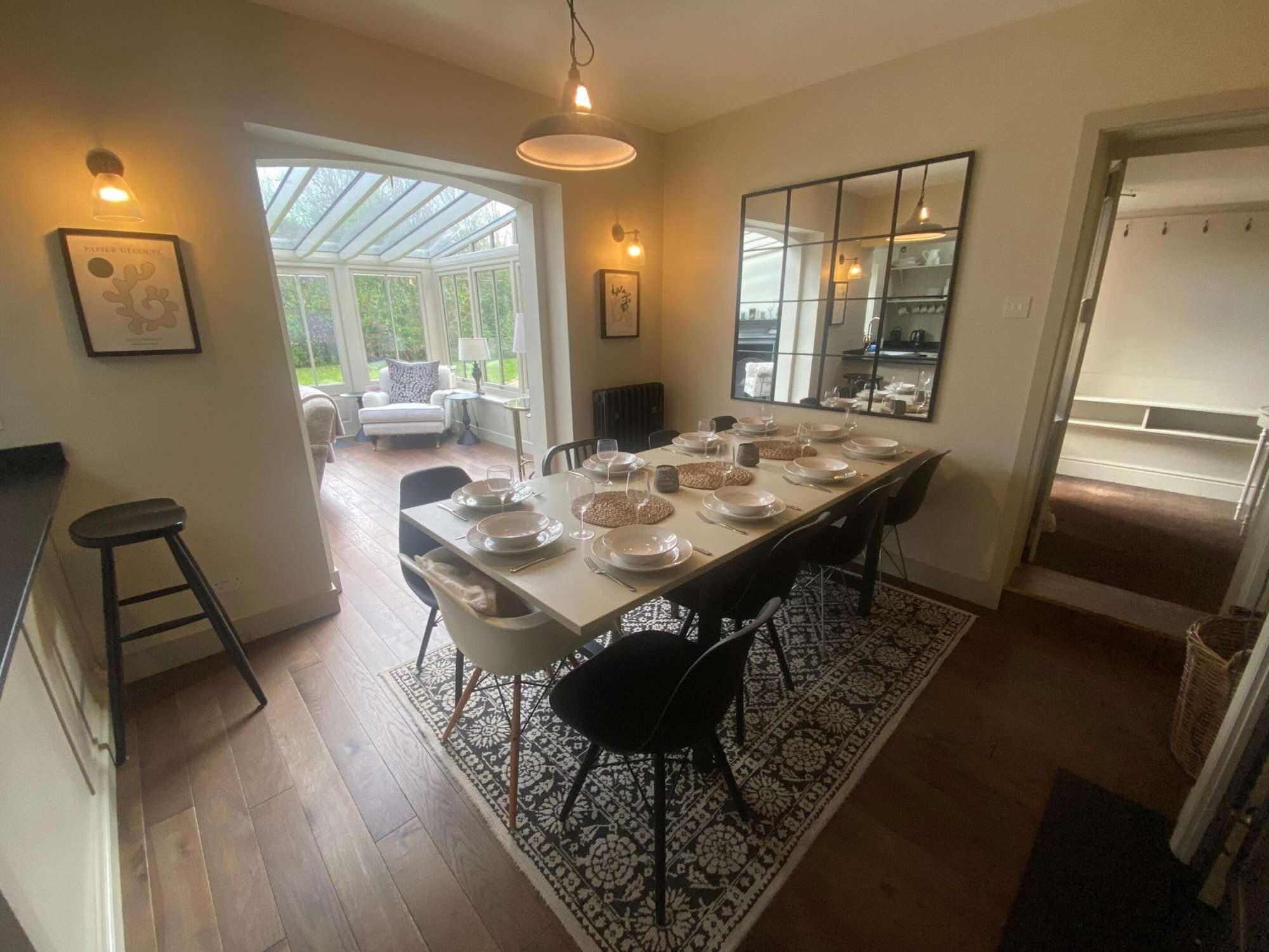 A dining area with a long table set for a meal, a kitchen in the background, and a conservatory with a view of greenery.