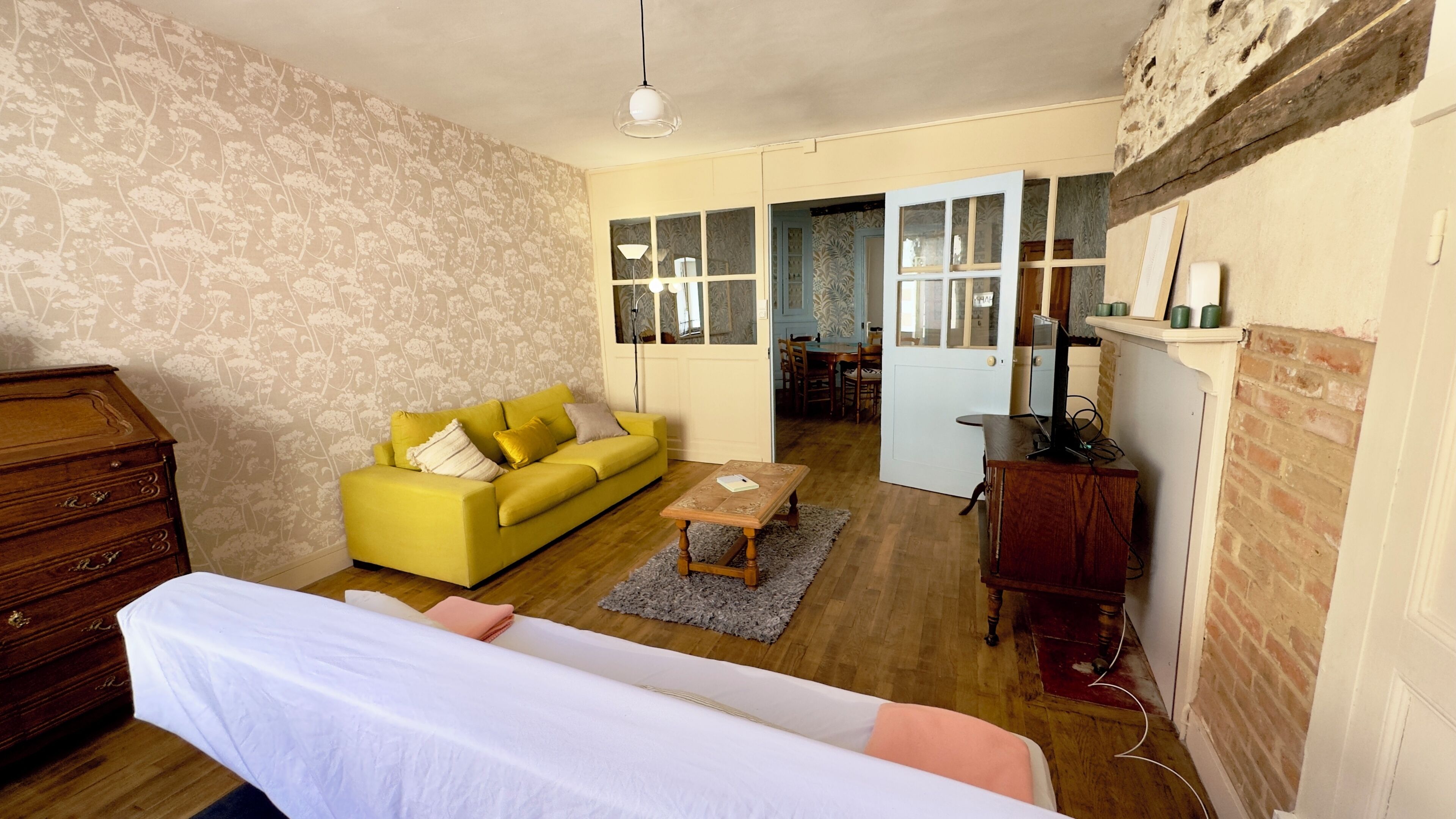 A room with a bed, a yellow sofa, a wooden dresser, and a dining area visible through an open door.