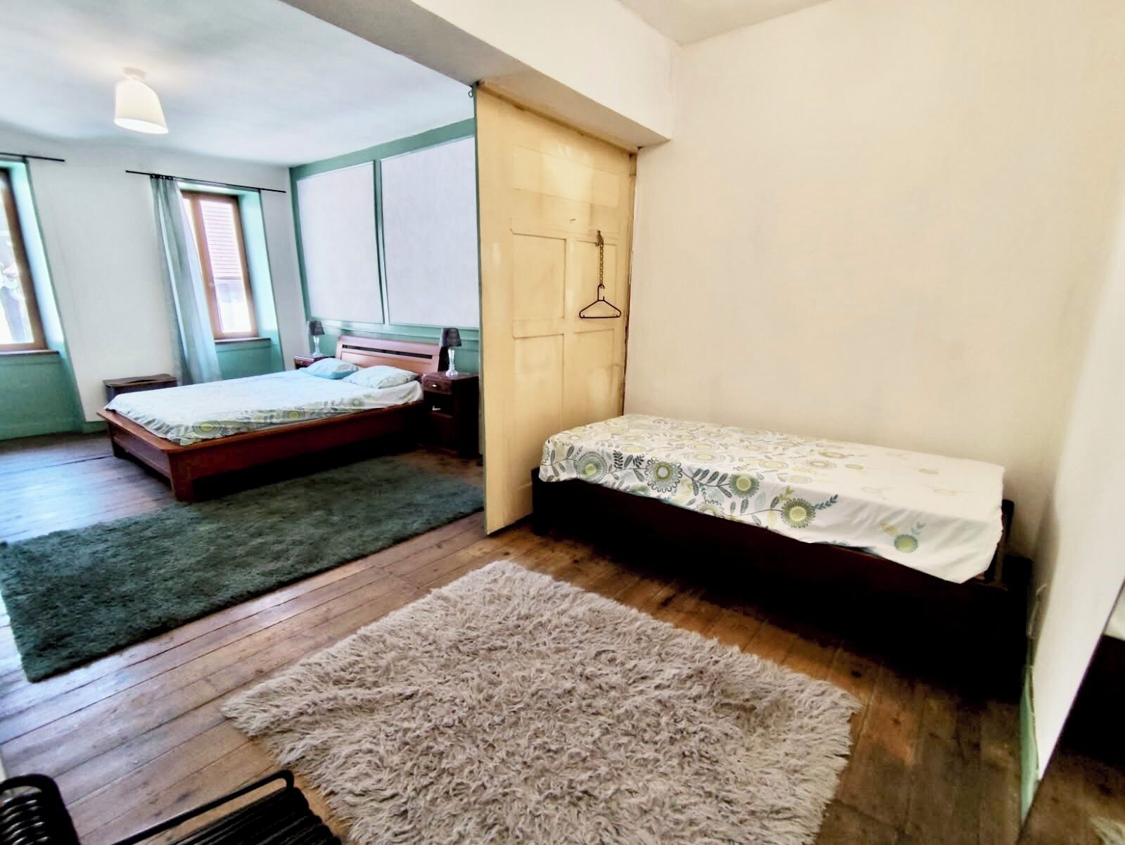 A room with a wooden floor, a large bed with a green rug, a smaller bed with a floral bedspread, a wooden wardrobe, and a hanging light fixture.