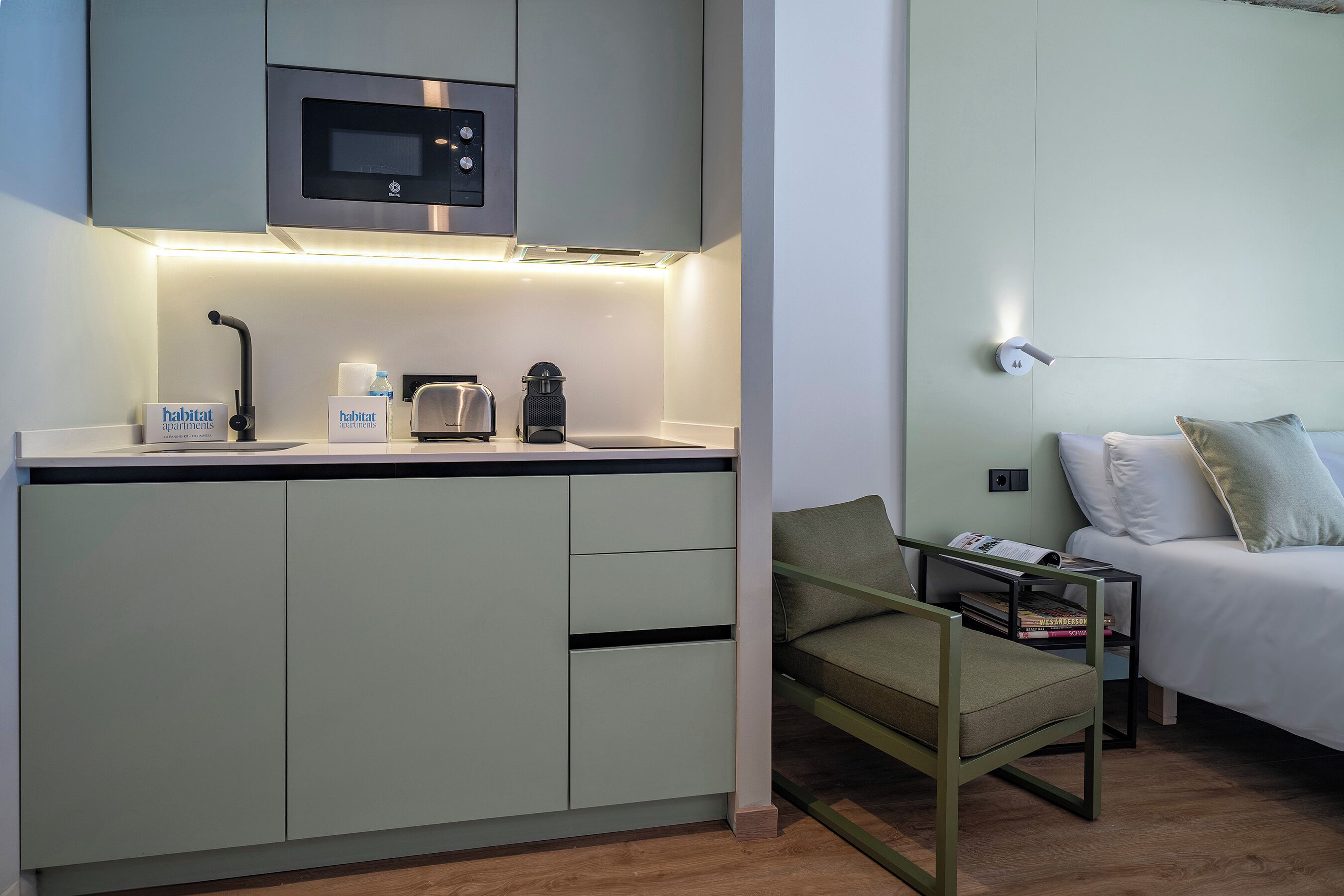 Et moderne tekøkken med mikrobølgeovn, brødrister og kaffemaskine. Der er et lille bord med et skilt for 'habitat apartments' og en stol. En seng med puder er synlig i baggrunden.