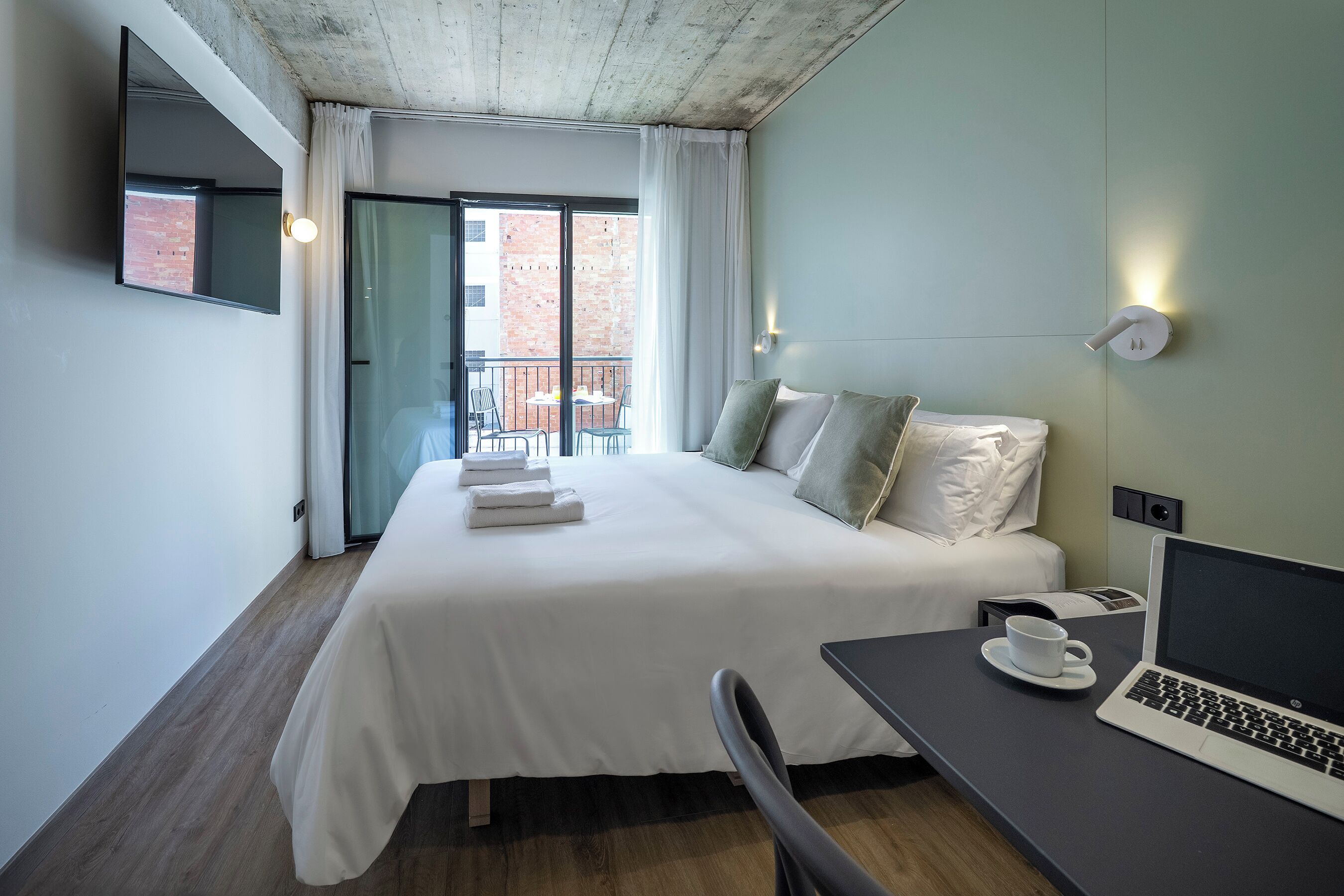 Et hotelværelse med seng, skrivebord med en bærbar computer, stol, et vindue med gardiner og udsigt til en murstensbygning.