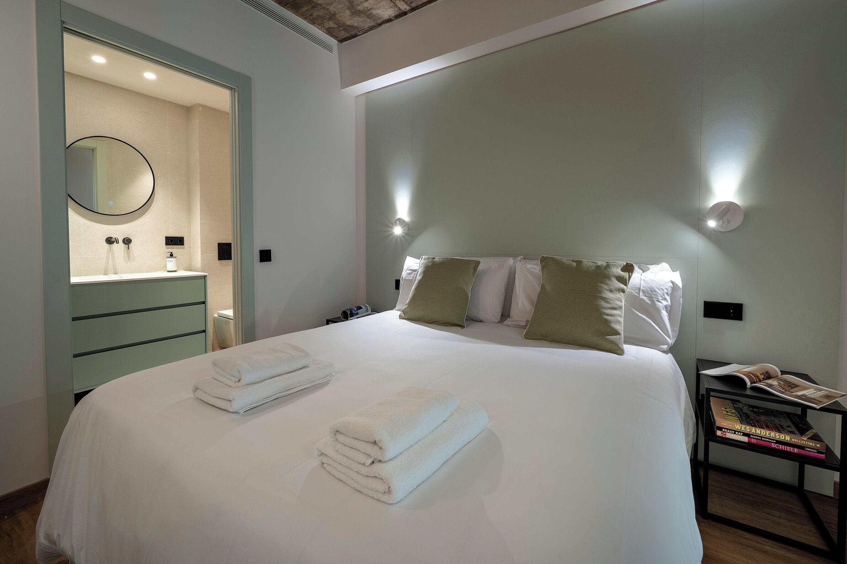 Cama arrumada com toalhas dobradas, mesa de cabeceira com uma revista e mesa lateral com abajur.