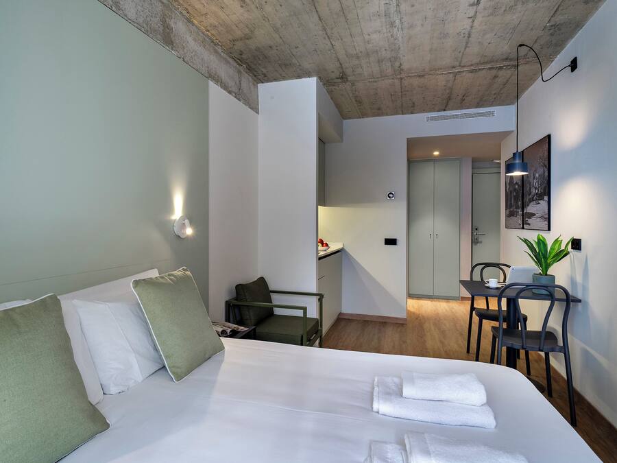 Habitación de hotel con cama, escritorio, silla, pequeña mesa, planta en maceta y espejo.