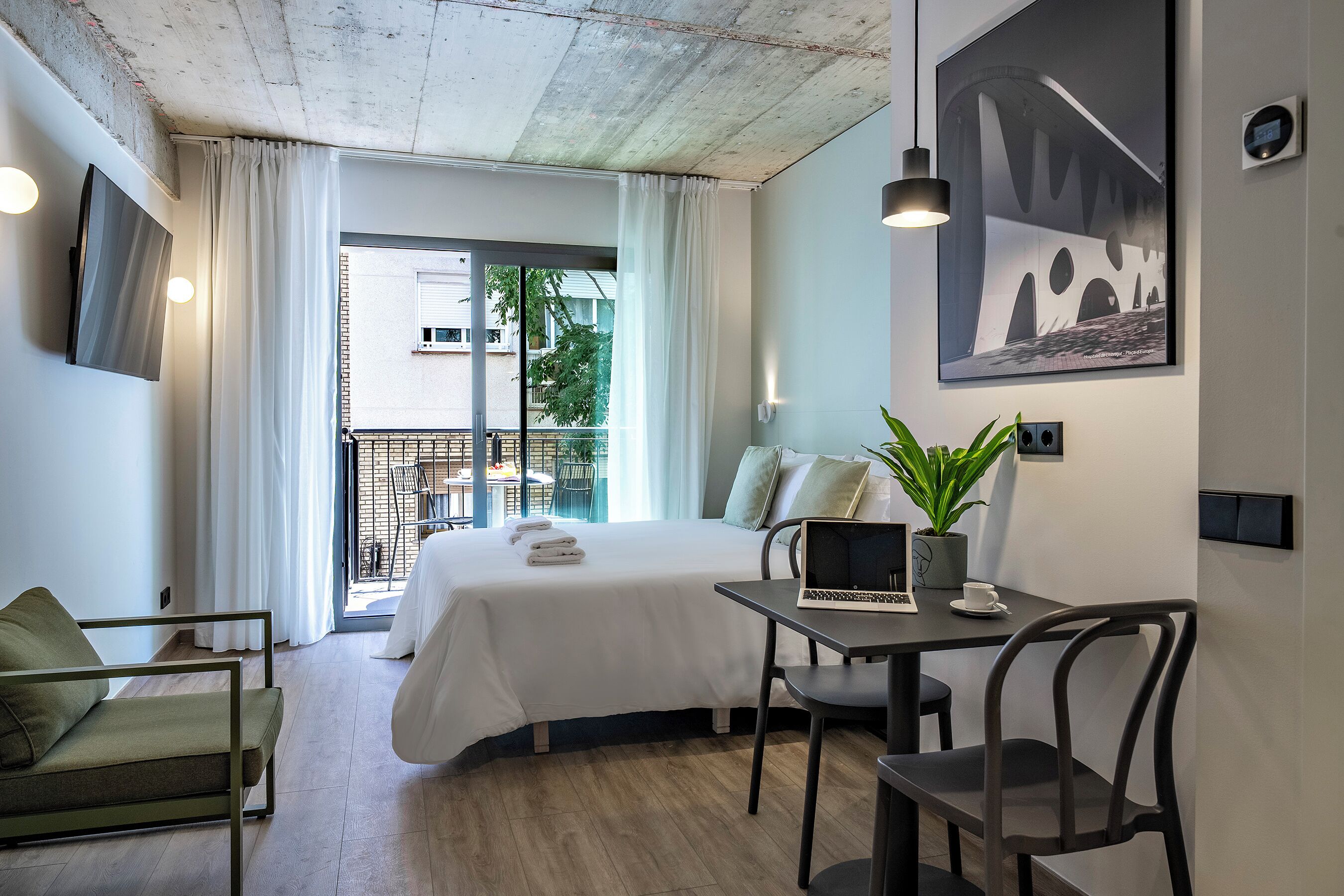 A hotel room with a bed, a desk with a laptop, a chair, a potted plant, and a view of the outside balcony.