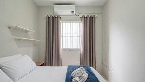 Ett hotellrum med en säng, ett fönster med gardiner, en väggmonterad luftkonditioneringsanläggning och en handduk på sängen.
