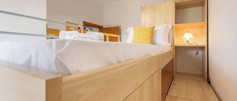 A compact room with a bed, a small desk with a lamp, and a wooden wardrobe.