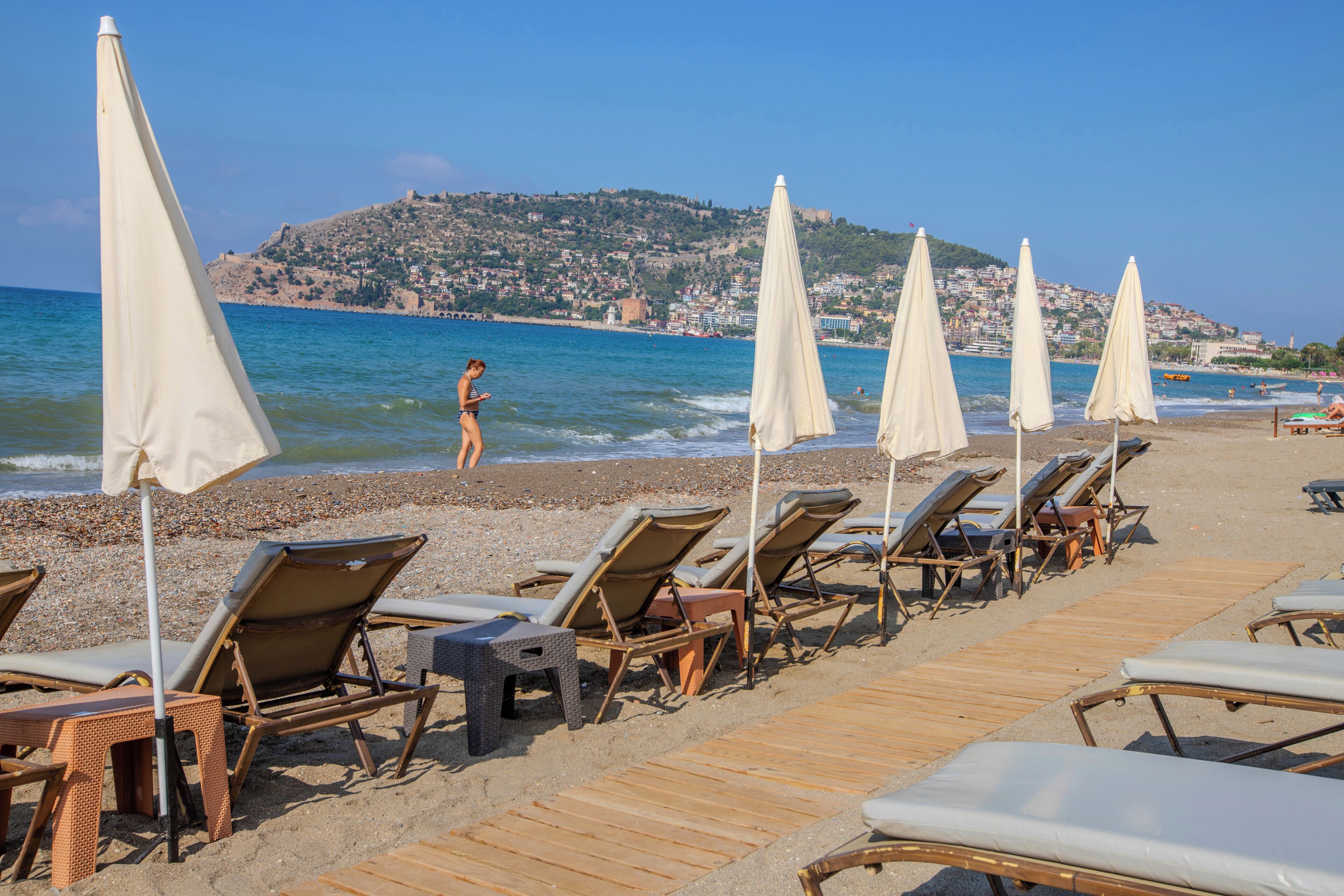 A beach with rows of sun loungers and umbrellas, a wooden walkway, and a view of the sea and a hillside town.