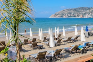 A beach with rows of sun loungers and umbrellas, a sign prohibiting food and drink on the beach, and a view of the sea and a distant hill. - Dublin Hotel  (Alanya)