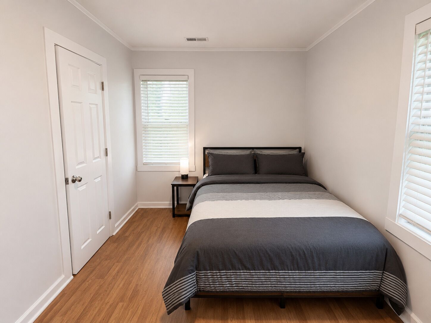Um quarto com uma cama, uma mesa de cabeceira, uma janela com persianas e uma porta.
