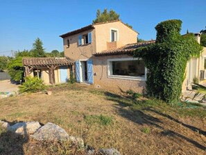 A two-story house with a tiled roof, a covered patio, and a garden area with dry grass and a rock. - A tropical getaway in a private villa near the sea and Nice on the French Riviera! (La Gaude)