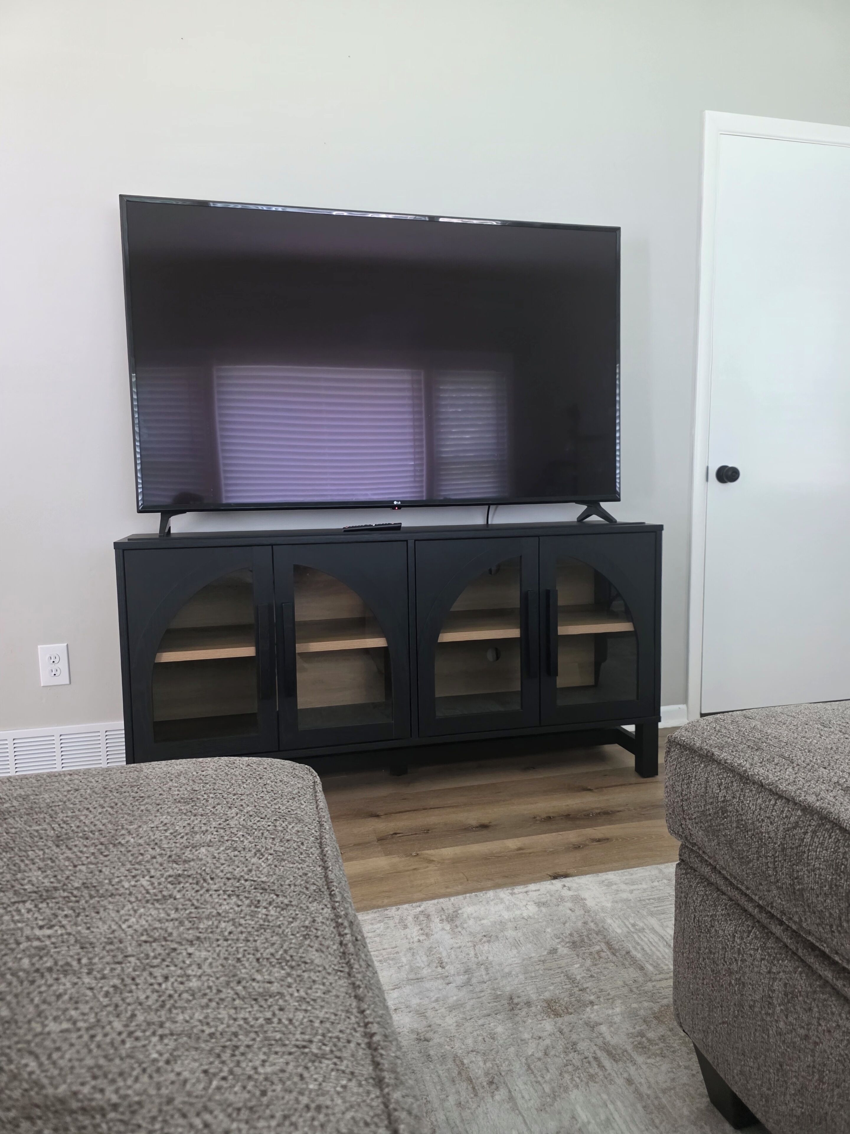 A room with a flat-screen TV mounted on a dark cabinet, a white door, and two grey sofas.