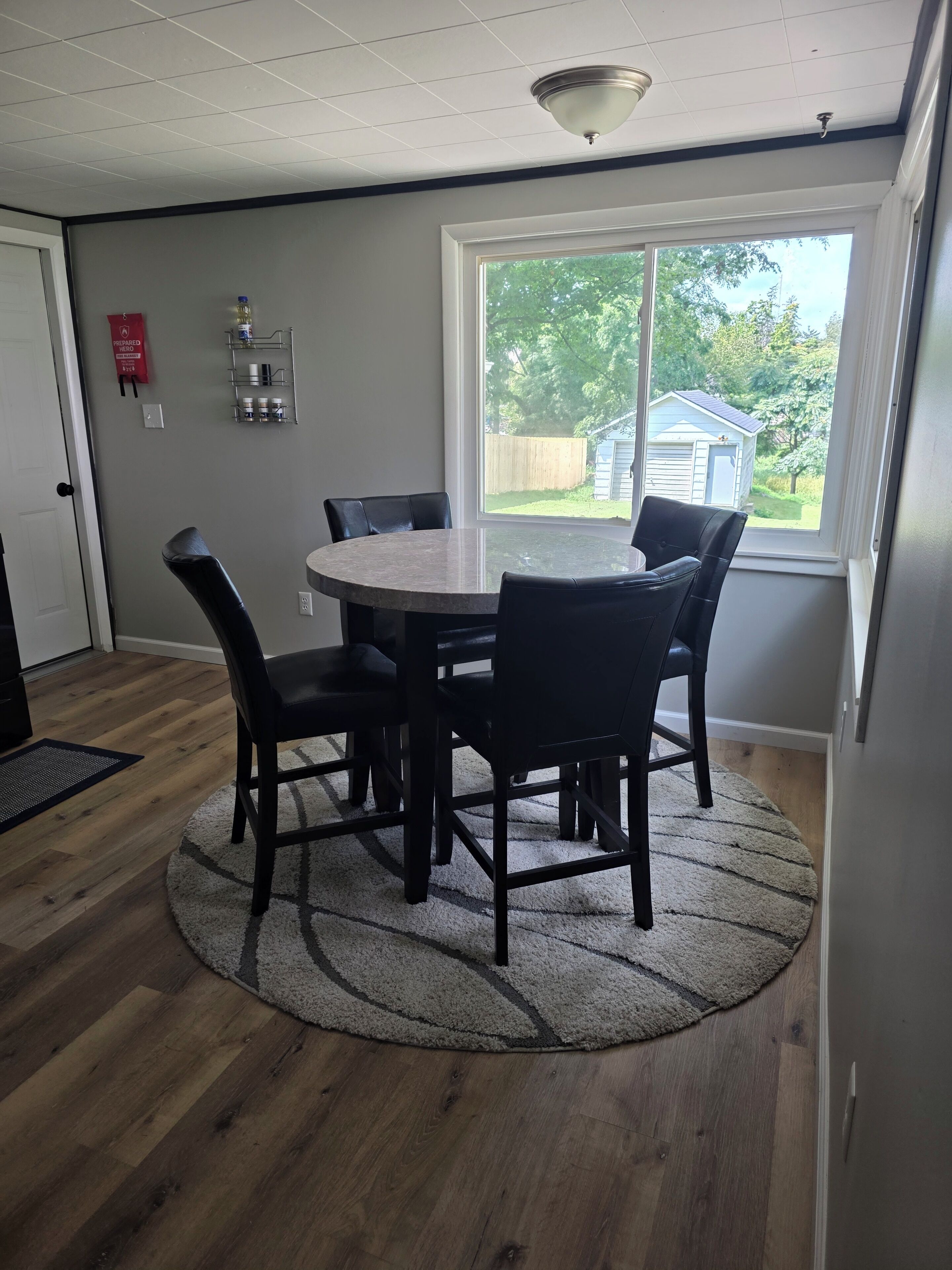Un coin repas avec une table ronde et des chaises, une grande fenêtre donnant sur l’extérieur et un extincteur fixé au mur.
