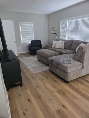 Ein Wohnzimmer mit einem Ecksofa, einem dunklen Sessel, einem Flachbildfernseher auf einem Standfuß und einer Stehlampe.