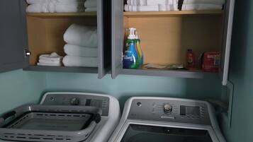 A laundry room with two washing machines and shelves above containing folded towels and cleaning supplies.