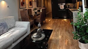 Een moderne woonkamer met een bank, een salontafel met decoratie, een eethoek met stoelen en een keuken met donkere kastjes.