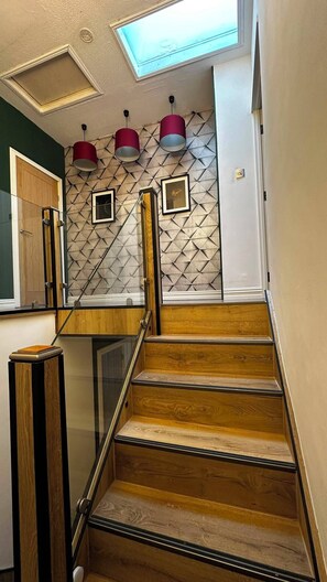 A staircase with wooden steps and glass railings, leading to a room with a geometric patterned wall and pendant lighting. - Modern Home in Surrey Quays/ Canada Water (London)