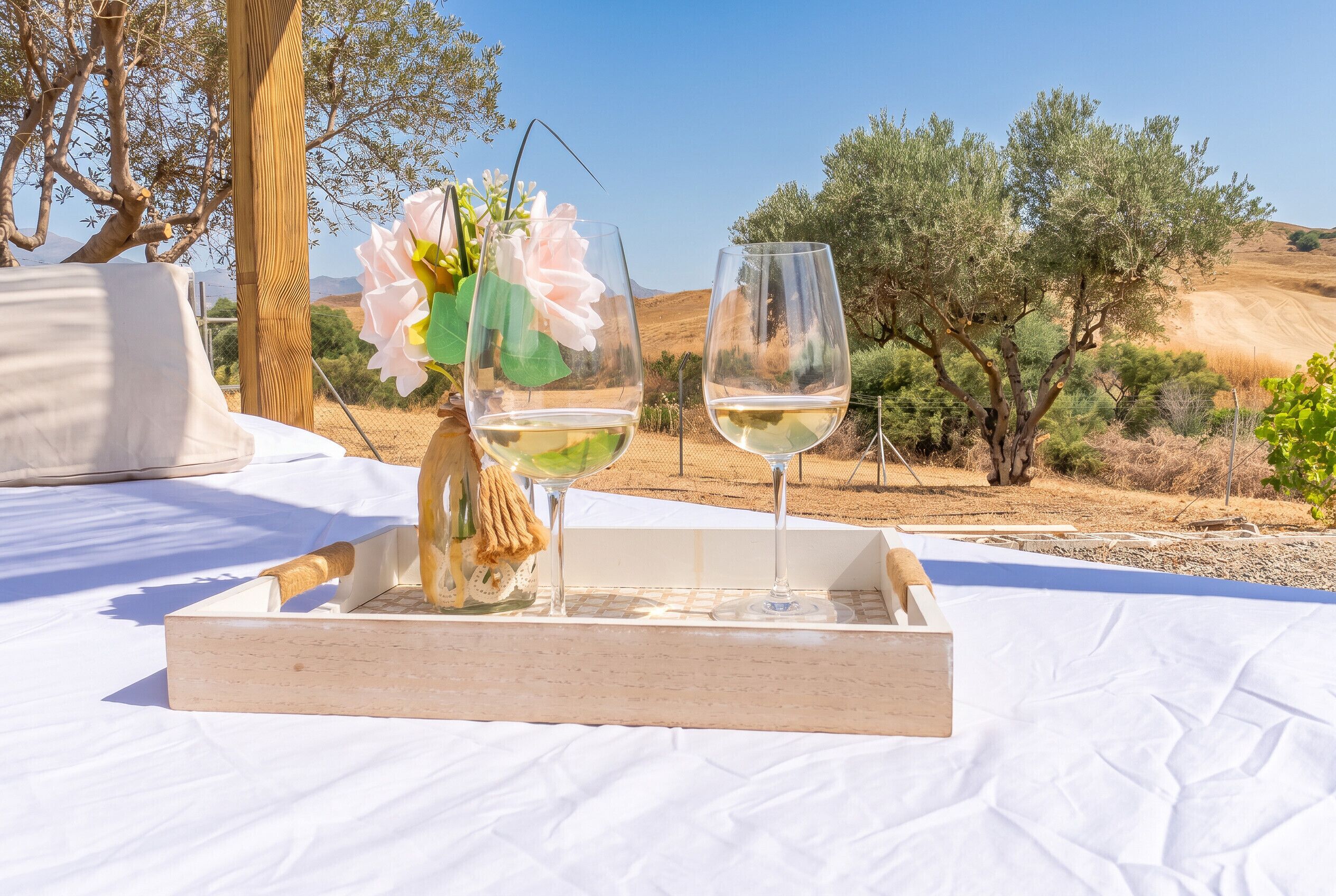 A wooden tray with two wine glasses and a vase with flowers on a white tablecloth.
