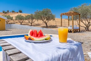 Une table placée au bord de la piscine, avec une assiette de tranches de pastèque et de mangue, un verre de jus d’orange et une terrasse en bois.