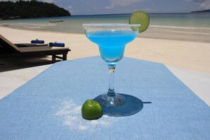 Een blauwe cocktail op een tafel, met een limoenpartje en een plakje limoen op een blauwe onderzetter. Achtergrond: een strand en de oceaan.