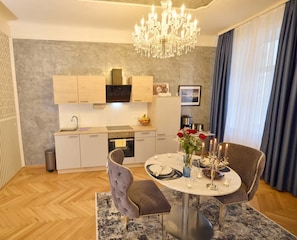 A modern dining area with a chandelier, a round table set for two, and a kitchen in the background. - Sophie's Place Augarten - Imperial Lifestyle Apartments (Wien)