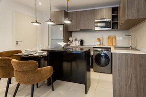 A modern kitchen with a central island, built-in appliances, and a dining area with chairs. - Luxe 1BR Apt at TheBantree, Airport (Accra)