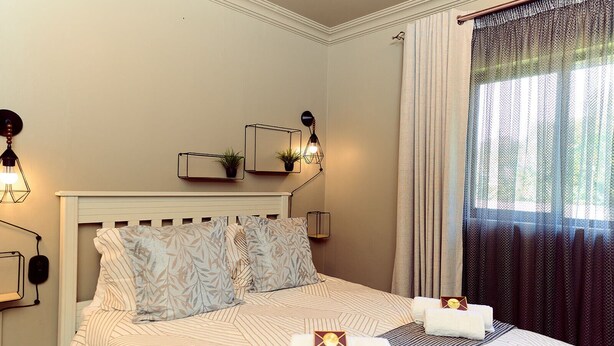 Une chambre d’hôtel avec un lit, deux tables de chevet, une lampe fixée au mur, une fenêtre avec des rideaux et une petite plante accrochée au mur.