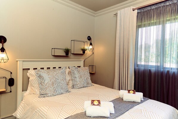 A hotel room with a bed, two bedside tables, a wall-mounted lamp, a window with curtains, and a small plant on the wall.