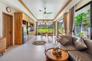 A modern kitchen with wooden cabinets, a dining area with a table and chairs, a sofa with cushions, and a view of a lush garden through large windows. - 2-bedroom house in peaceful Gianyar with private pool and natural views (Gianyar)