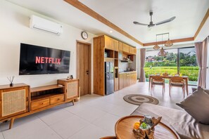 A modern living room with a flat-screen TV, wooden furniture, a dining area, and a view of a lush garden. - 2-bedroom house in peaceful Gianyar with private pool and natural views (Gianyar)