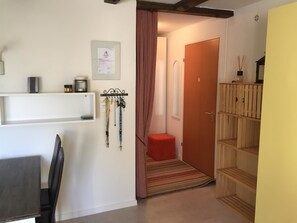 Een kleine kamer met een bed, een tafeltje en een stoel. Er is een deur naar een andere kamer en een plank met spullen erop.