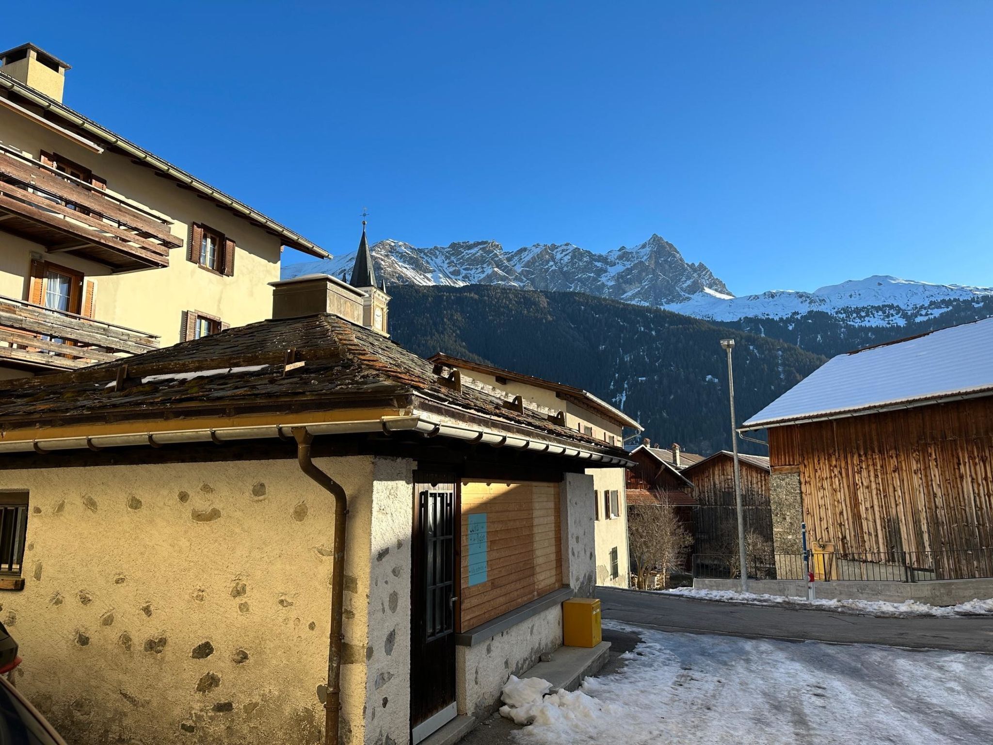 Un village de montagne traditionnel, avec des bâtiments rustiques, une clôture en bois et une vue dégagée sur les montagnes enneigées qui se dressent à l’horizon.
