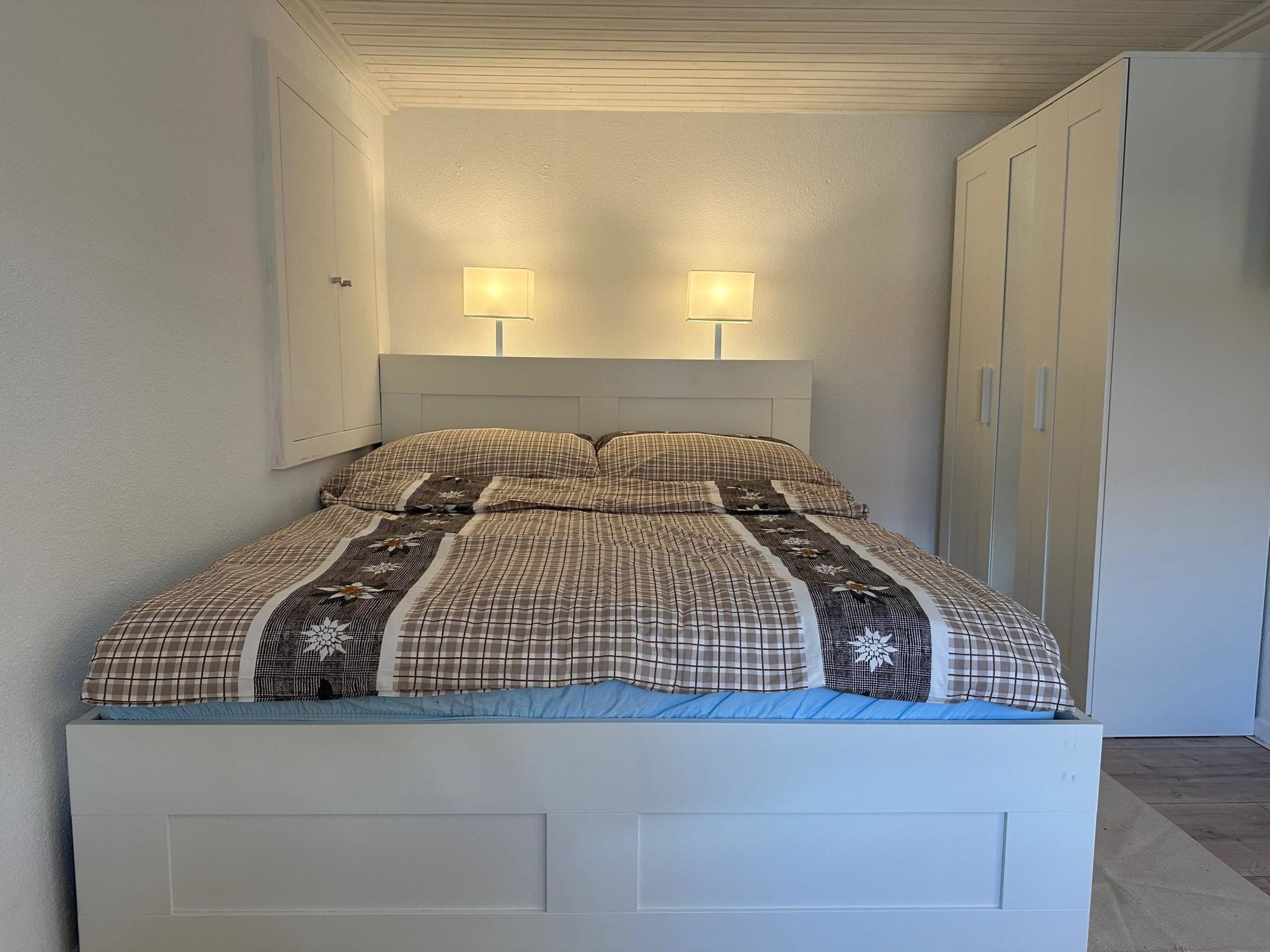 A single-bed room with a white headboard, two bedside lamps, and a wardrobe with a door.