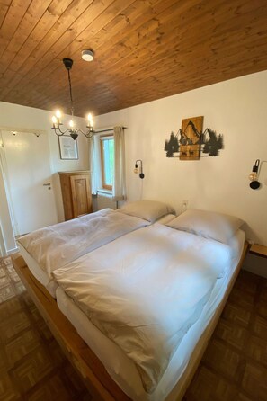 A bedroom with a wooden ceiling, a bed with white bedding, a window with curtains, and a wall-mounted light fixture. - Tga Muntanela Appartement/Fewo, Dusche und Bad, WC, 2 Schlafräume by Interhome (Savognin)