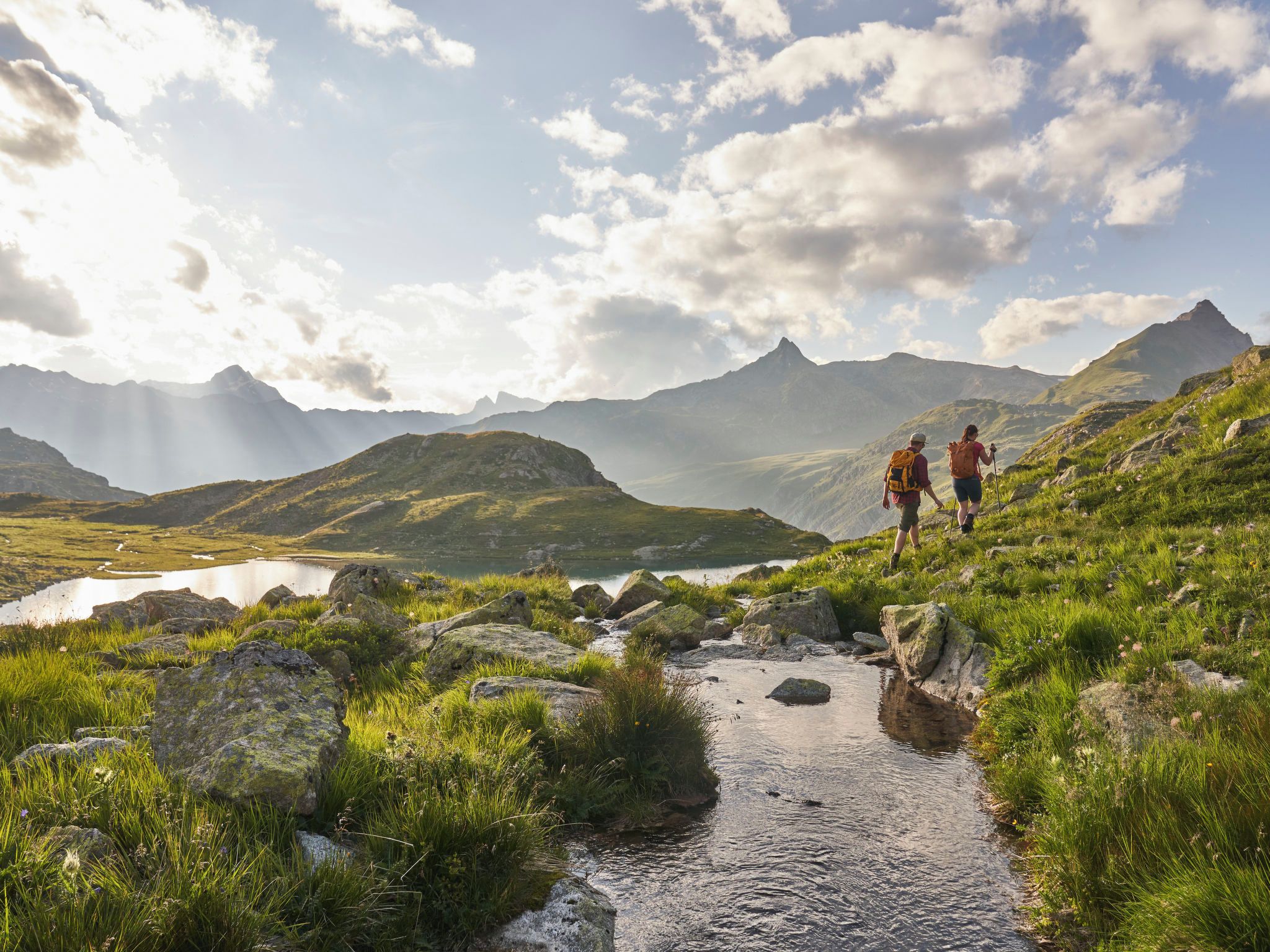 Vuoristoinen maisema, jossa on patikoijia, puro ja järvi.