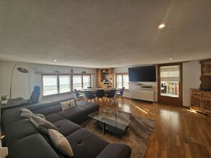 A modern living room with a sectional sofa, a dining area with a table and chairs, a kitchen with a TV, and a large window. - Bavegna Appartement/Fewo, Dusche und Bad, WC, 3 Schlafräume by Interhome (Savognin)