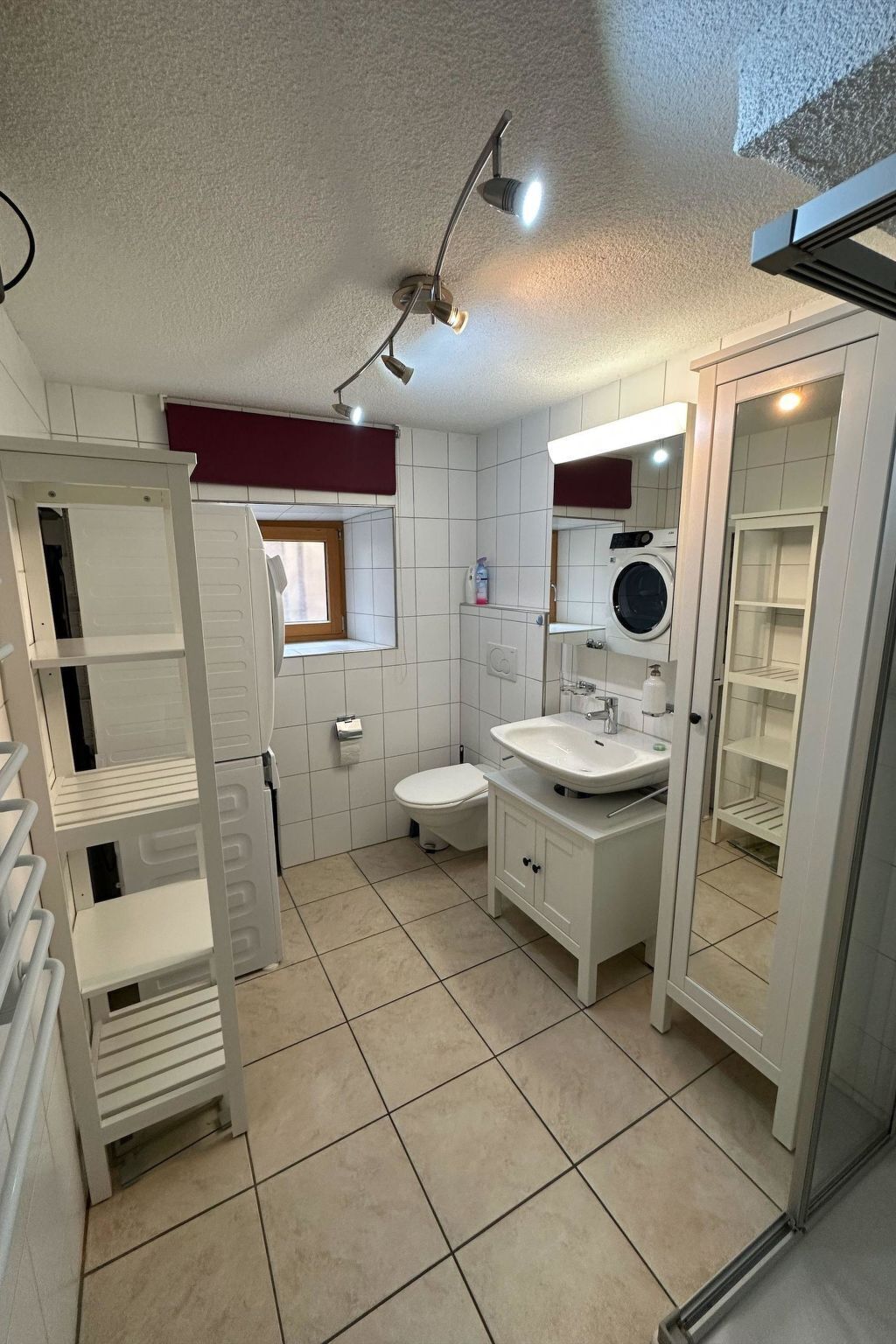 Baño con lavamanos blanco, inodoro y lavadora. Hay un espejo sobre el lavamanos y un estante con artículos de aseo.