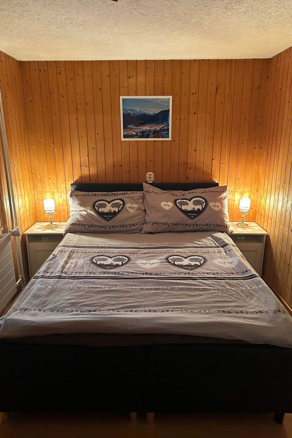 A bed with a patterned bedspread and heart-shaped pillows, flanked by two bedside lamps, in a room with wooden paneling and a framed picture on the wall.