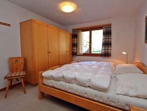 A bedroom with a wooden bed, a wooden wardrobe, a chair, and a window with curtains. - Soras 10 A Appartement/Fewo, Bad, WC, 2 Schlafräume 3 Zimmerwohnung - 4 Betten by Interhome (Savognin)
