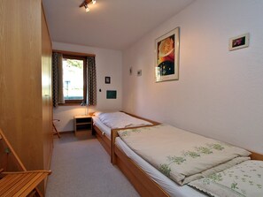 Ein kleines Zimmer mit einem Einzelbett, einem Holzbett, einem Nachttisch, einem Stuhl, einem Fenster mit Vorhängen und einem an der Wand hängenden Bild.