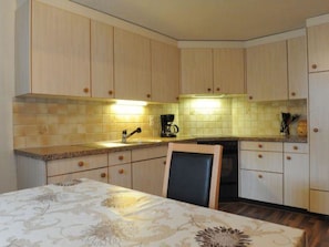 A kitchen with wooden cabinets, a tiled backsplash, a coffee maker, and a table with a patterned tablecloth. - Ferienwohnung Hof Collet Appartement/Fewo, Dusche oder Bad, WC, 1 Schlafraum by Interhome (Cunter)