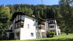 A two-story building with balconies, a grassy lawn, and a forested hillside in the background. - Soras Zerbe/Hirth Appartement/Fewo, Bad, WC, 1 Schlafraum by Interhome (Savognin)