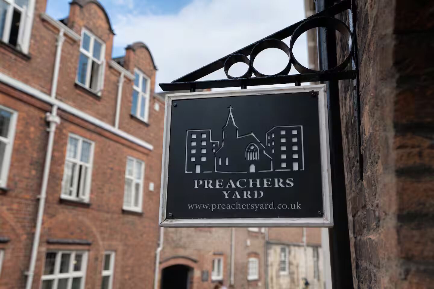 Letrero que dice 'Preachers Yard' con una dirección web, sobre un fondo de edificios de ladrillo rojo.