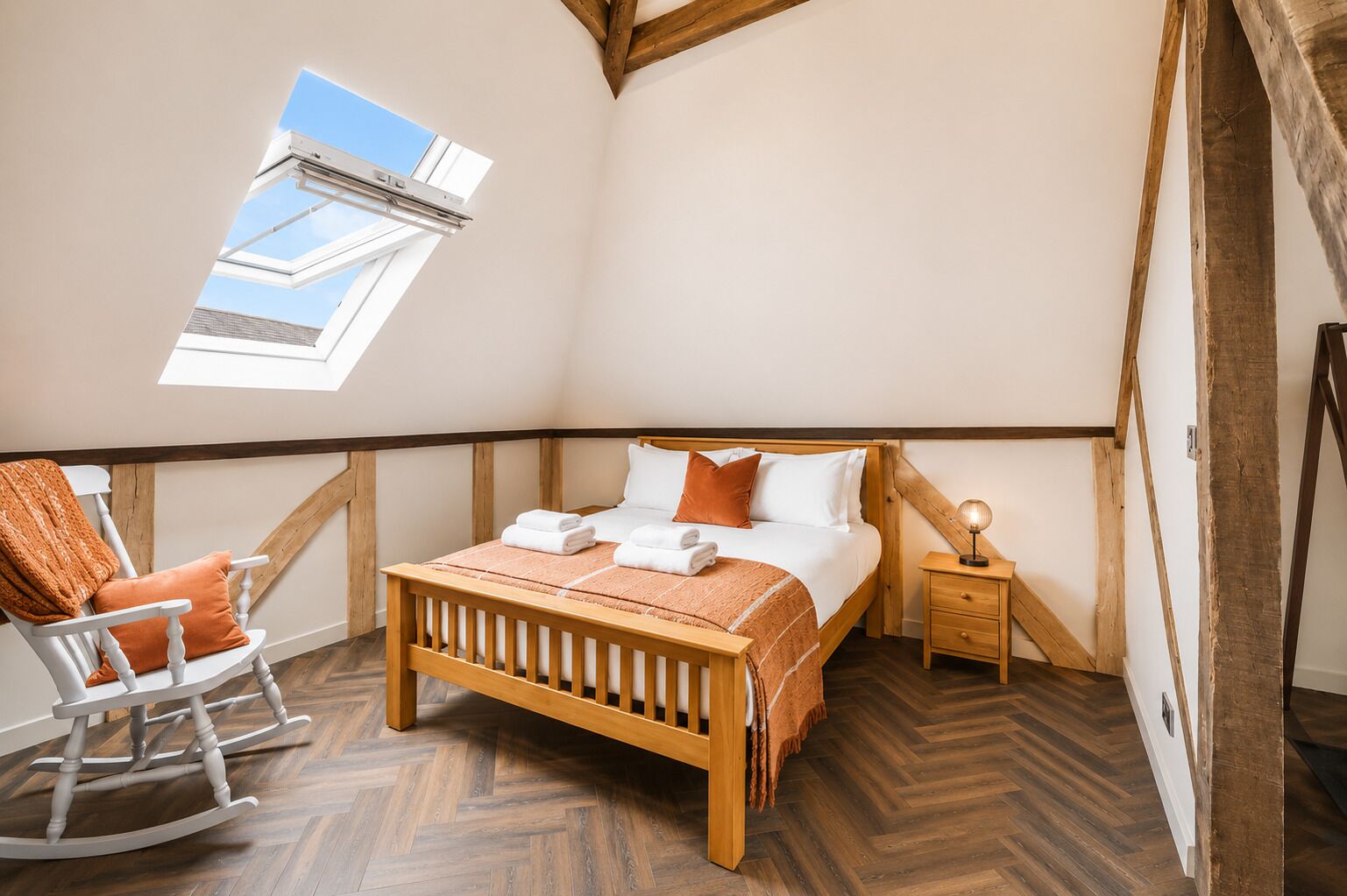 Quarto com teto inclinado, cama de madeira, cadeira de balanço, mesa de cabeceira e claraboia.